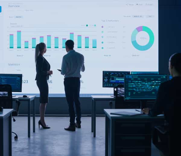 Co-workers standing in front of a large screen showing a dashboard of banking information, in a control room.